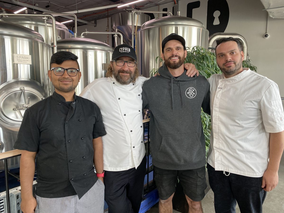 four men pose for photo in front of brew tanks