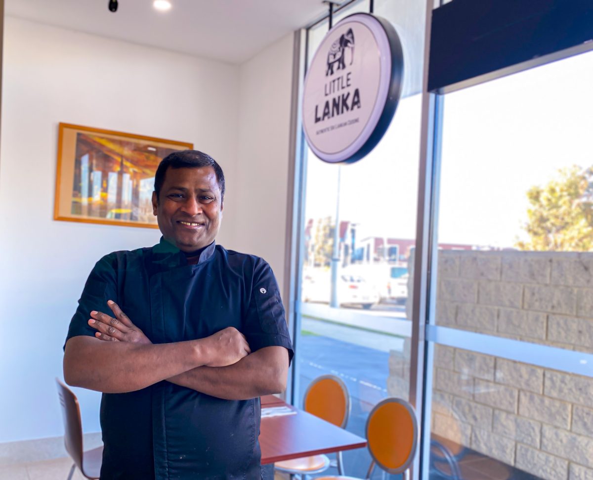 Nelson wears a black chefs jacket and smiles in front of Little Lanka sign