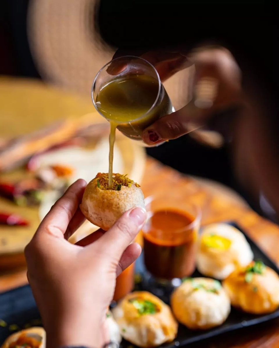 One hand holds a small 'pani puri' ball while the other pours a green liquid into it. A plate of pani puri is in the background