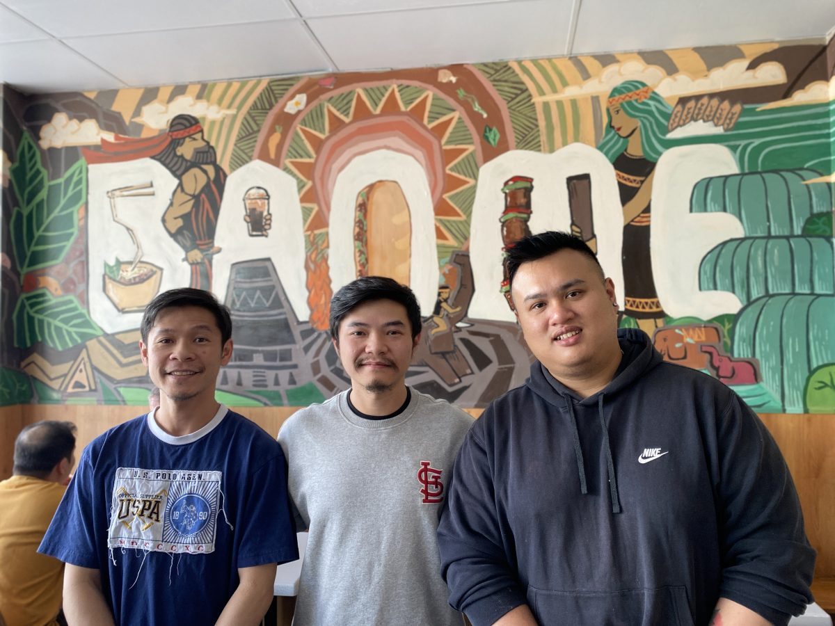 Three Vietnamese men stand in front of a mural in a restaurant.